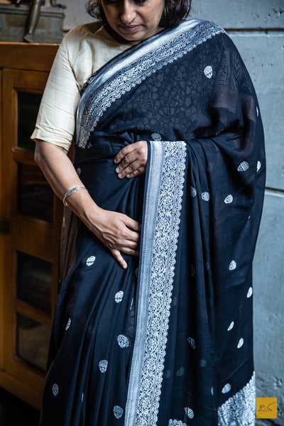 Make a statement in this black beauty Lavanya Banarasi Georgette Handwoven Saree. Exquisitely crafted with silver zari kadhwa motifs, this luxuriously soft saree is designed to perfectly drape for a beautiful silhouette. Ideal for both formal and informal occasions, this timeless piece of art is sure to turn heads.  Fabric- Georgette