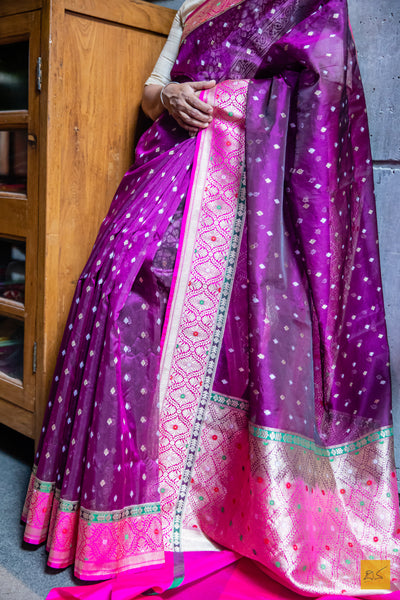 This NANDITHA banarasi kora silk organza saree is a luxurious piece of art. Pristinely handwoven and detailed with a stunning meenakari border and pallu, it is a sheer elegance for any special occasion. Its intricate small motifs add a delicate beauty, making it an effortlessly chic and eye-catching piece. Gracefully drape it around and be the centre of attention.  Fabric- Kora silk Organza  Colour- Purple and Pink